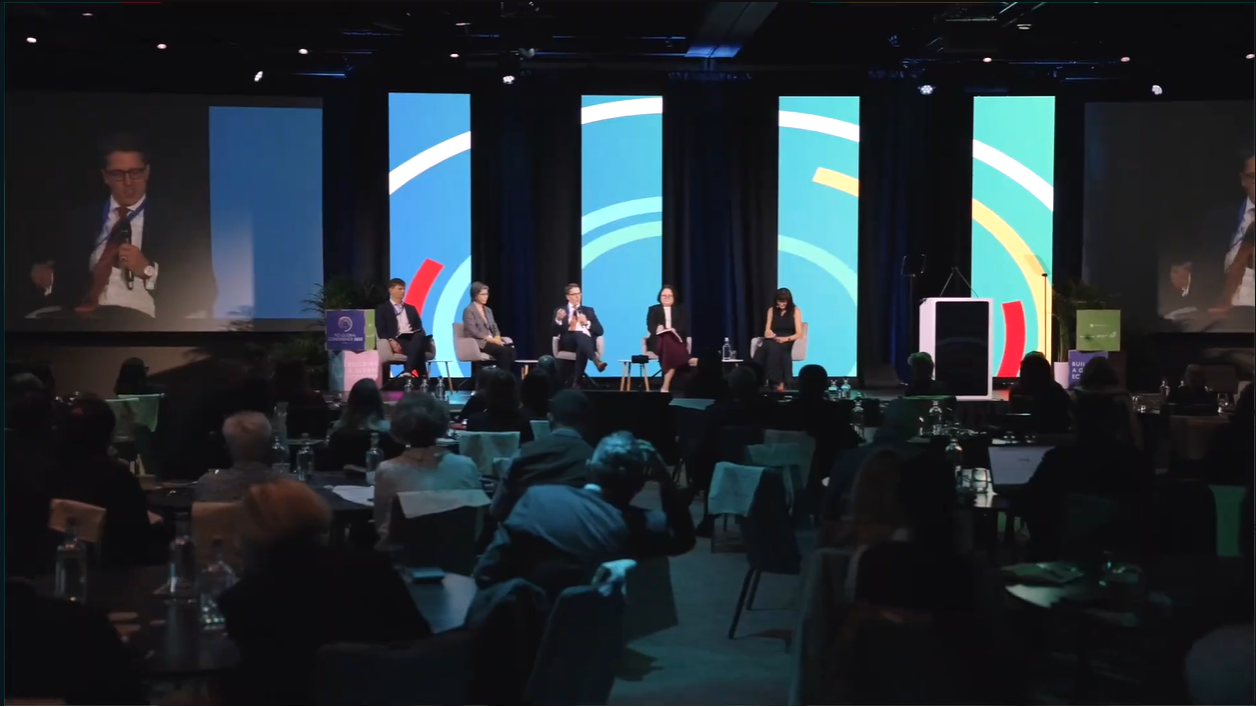 A panel of 5 speakers on stage in a dark conference hall